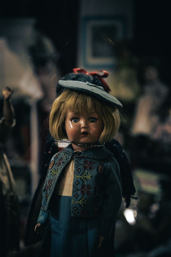 a slightly creepy procelain doll with a blond pageboy haircut, blue eyes, a grey hat, and a blue suit-dress.